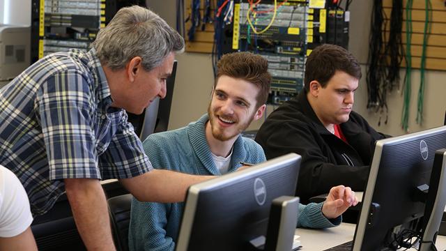 2名微笑的学生在电脑前工作，一名教师俯身帮助他们