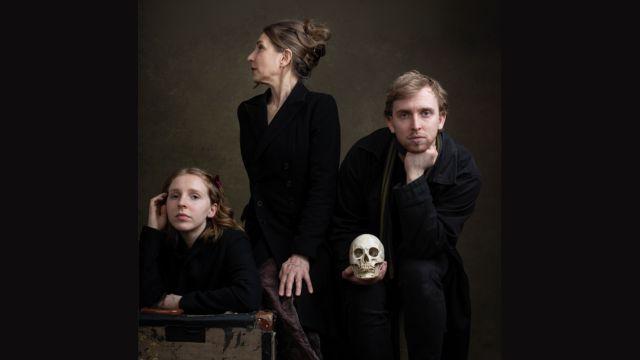 Three actors in black pose with a skull for a studio portrait against a dark backdrop.
