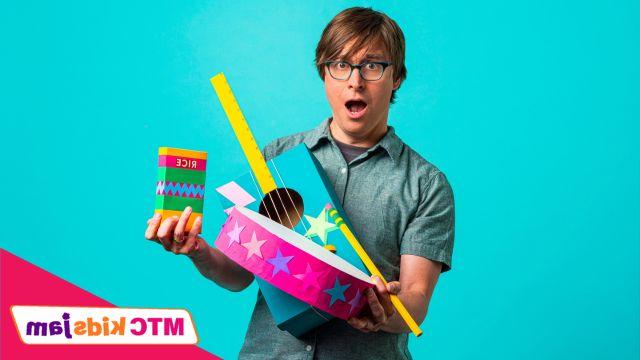 Justin Roberts holding brightly colored paper crafts in the shapes of instruments. He is standing against a bright blue background.
